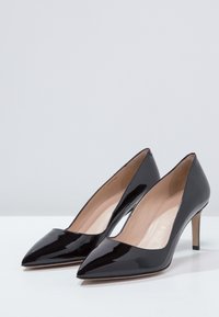 Černé lakované lodičky s špičatou špičkou a béžovou vnitřní výplní, vystavené vedle sebe na bílém povrchu.