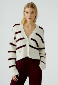 Gestrickte, verkürzte Strickjacke in Creme mit kastanienbraunen horizontalen Streifen. Sie verfügt über einen tiefen V-Ausschnitt, zwei Fronttaschen und perlähnliche Knöpfe.