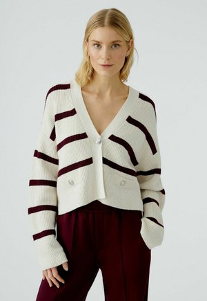 Gestrickte, verkürzte Strickjacke in Creme mit kastanienbraunen horizontalen Streifen. Sie verfügt über einen tiefen V-Ausschnitt, zwei Fronttaschen und perlähnliche Knöpfe.