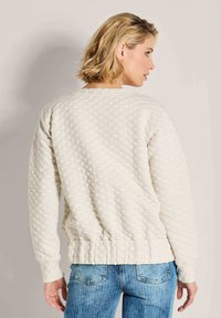 Vrouw met kort blond haar, gekleed in een gewatteerde off-white trui en blauwe jeans, staand en kijkend naar haar rechterzijde tegen een effen achtergrond.