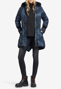 Quilted blå camouflagejakke med hætte, struktureret stof, frontlynlås og sidelommer lagdelt over en mørk sweater og sorte jeans.