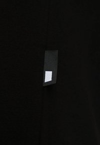 Tissu noir avec une petite étiquette rectangulaire noire présentant une forme géométrique blanche cousue dans la couture.