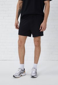 Zwarte sportshorts van zacht materiaal, voorzien van een elastische tailleband en zijzakken. Draagt witgekleurde sokken en zilveren hardloopschoenen.