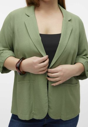 Femme ajustant un blazer vert clair sur un haut noir, portant un bracelet en écaille et un jean bleu foncé, sur fond blanc.