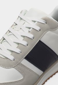 Baskets blanches avec accents en daim beige, bande latérale en cuir noir et lacets blancs sur une semelle blanche.