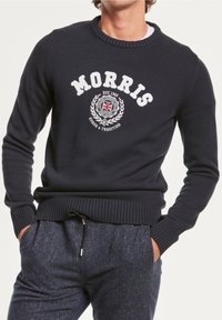 Homme portant un pull bleu marine avec le texte "MORRIS" et un emblème du Royaume-Uni, associé à un pantalon bleu texturé, les mains dans les poches.