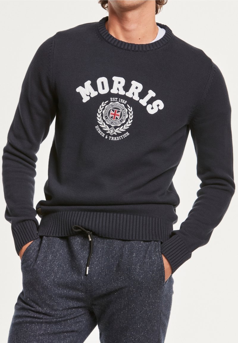 Homme portant un pull bleu marine avec le texte "MORRIS" et un emblème du Royaume-Uni, associé à un pantalon bleu texturé, les mains dans les poches.
