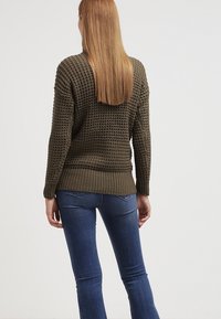 Suéter tejido de color verde caqui con patrón texturizado, de corte holgado y ribete acanalado; llevado sobre jeans de denim azules. Longitud y diseño estándar.