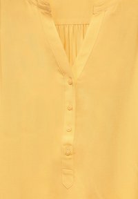 Gul blus med V-ringning, plisserad frontdetalj och sex knappar ner längs knappslån. Slätt tyg med en mjuk textur.