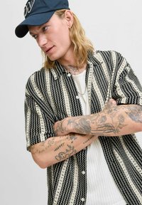 Camisa de manga corta con un patrón en blanco y negro, con botones visibles y un diseño texturizado. Llevada sobre una camiseta blanca con canalé.