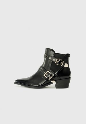 Black ankle boot made of glossy leather, featuring pointed toe, black and silver buckles, and a low heel with a textured sole.