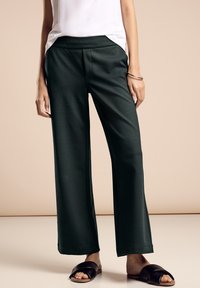Pantaloni a gamba larga di colore verde scuro realizzati in tessuto morbido, dotati di una vita elastica e tasche laterali, abbinati a sandali neri a ciabatta.