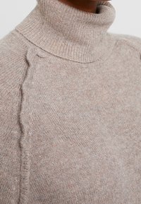 Pull à col roulé beige clair en tissu tricoté, avec col côtelé et texture subtile offrant une apparence douce et chaleureuse.