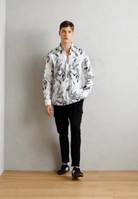 Hvit skjorte med svart bladmønster, paret med svarte bukser og svarte sneakers, stående på tregulv mot en grå vegg.
