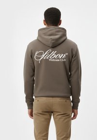 Sudadera marrón con un bolsillo canguro frontal, que presenta un gran logotipo blanco de "Silbon Wellness Club" en la parte posterior. Combinada con pantalones beiges.