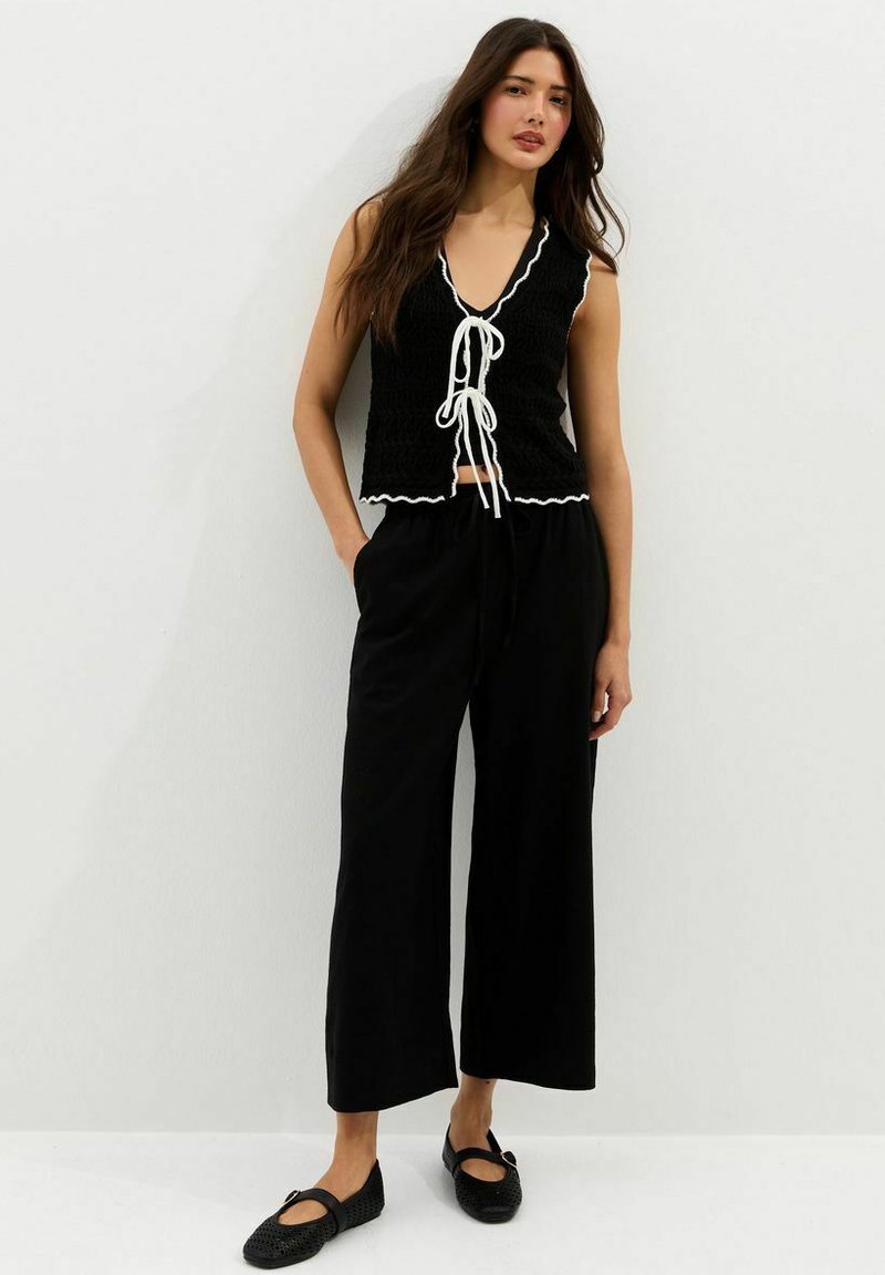 Woman standing against white wall wearing sleeveless black top with white ties, wide-leg black pants, and black woven flats.