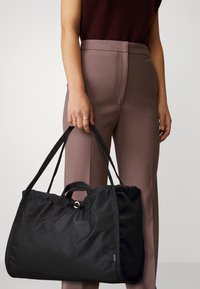 Femme tenant un grand sac fourre-tout en nylon noir, portant un pantalon taille haute mauve et un top sans manches bordeaux, avec des bagues dorées aux doigts.
