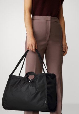 Femme tenant un grand sac fourre-tout en nylon noir, portant un pantalon taille haute mauve et un top sans manches bordeaux, avec des bagues dorées aux doigts.