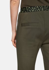 Dark green trousers with a textured black and green leopard print waistband and a single back pocket, showcasing a smooth fabric finish.