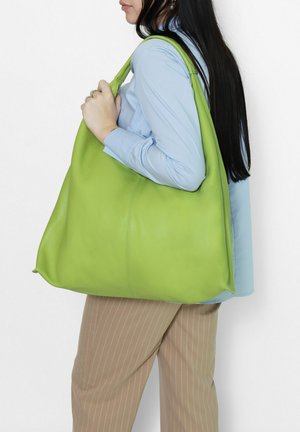 Woman wearing a light blue shirt and beige pinstripe pants carrying a large lime green leather shoulder bag on her right shoulder.