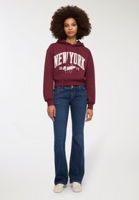 Burgundowy krótki hoodie z białym nadrukiem "New York", w połączeniu z ciemnoniebieskimi dzwonami i beżowymi sneakersami. Gładki materiał i luźny krój.