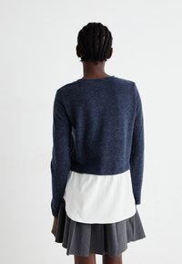Pull en maille bleu marine avec un design court, doté d'un ourlet superposé blanc. Le modèle l'associe à une jupe plissée grise, vue de dos.