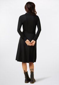 Abito lungo nero a maniche lunghe con vita stretta, gonna a campana e scollatura alta. Indossato con stivali neri alla caviglia; mani intrecciate dietro la schiena.