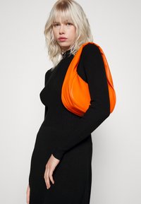 Sac en cuir orange avec une forme douce et arrondie, porté sur l'épaule. Robe noire à manches longues côtelées, au style minimaliste, sur fond uni.