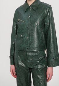 Zielona, krótkia kurtka z ekologicznej skóry o gładkiej, fakturowanej powierzchni, z zatrzaskami i kieszenią na piersi zapinaną na zamek.