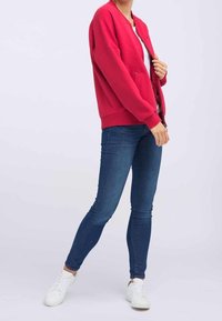 Sweatshirt rouge à manches longues avec poignets côtelés, porté par-dessus une chemise blanche. Associé à un jean skinny bleu et des baskets blanches.