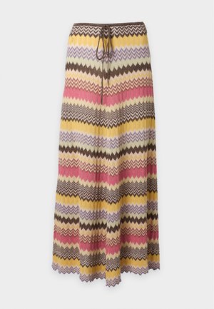 Femme portant un chemisier blanc sans manches froncé et une longue jupe multicolore à motif zigzag avec des sandales à talons noires, debout sur fond blanc.