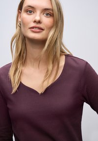 Donna bionda con occhi verdi che indossa piccoli orecchini a cerchio e un top ribbed di un viola scuro con scollatura a V, su uno sfondo chiaro e semplice.