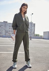 Olive green zip-up jacket and matching pants made of soft fabric, with relaxed fit and cuffed ankles. Person holding a drink, wearing light sneakers.