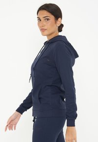 Felpa con cappuccio color navy, con zip, cordino e tasche laterali. Presenta polsini a coste e un piccolo logo sul petto.