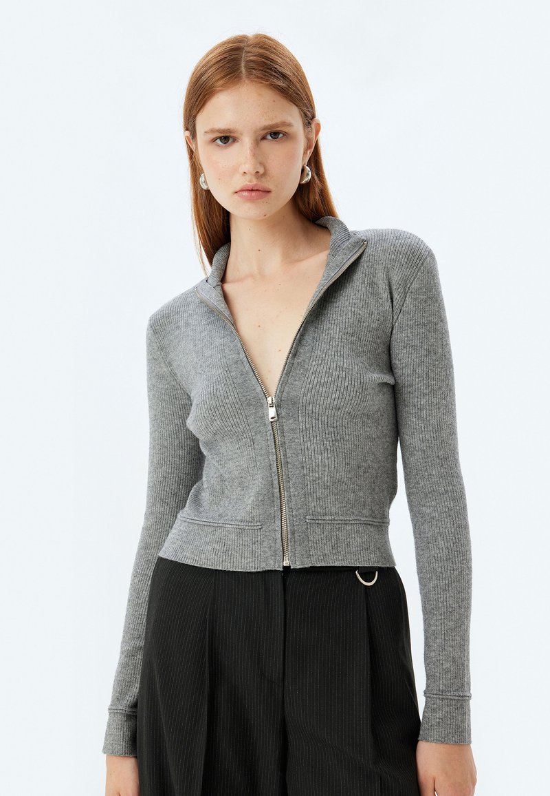 Cardigan grigio con zip, texture a coste, colletto alto e silhouette aderente. Presenta una zip argentata e elementi di design minimalisti.