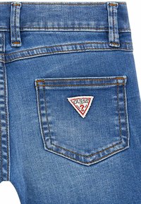 Jeans in denim blu con una toppa triangolare del logo Guess sulla tasca posteriore destra. Presentano cuciture marroni e un classico design a cinque tasche.