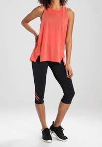 Top atletico senza maniche color corallo con fessure laterali, abbinato a leggings neri 3/4 con accenti rosa. Le scarpe da ginnastica nere completano il look.