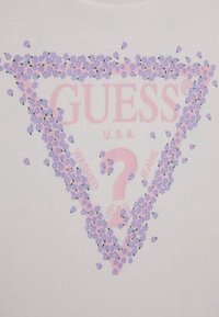 Valkoinen t-paita, jossa on pinkki ja violetti kukkatrikoon kolmio graafinen kuva ja "GUESS U.S.A." teksti. Pehmeä kangas, jolla on sileä rakenne.