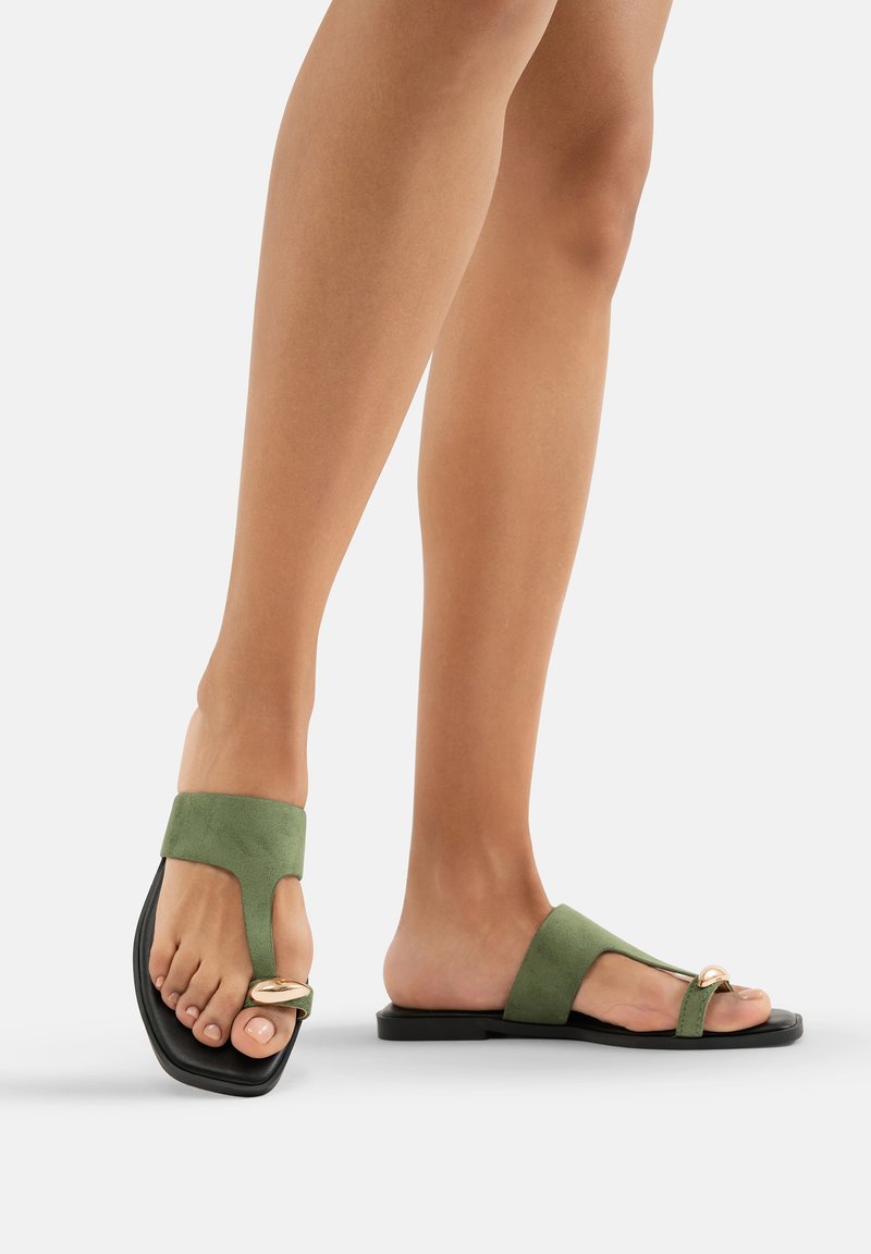 Sandales en suède vert avec une boucle au niveau des orteils et des sangles larges, dotées d'une semelle noire lisse et d'un détail doré. Conçues pour le confort et le style.