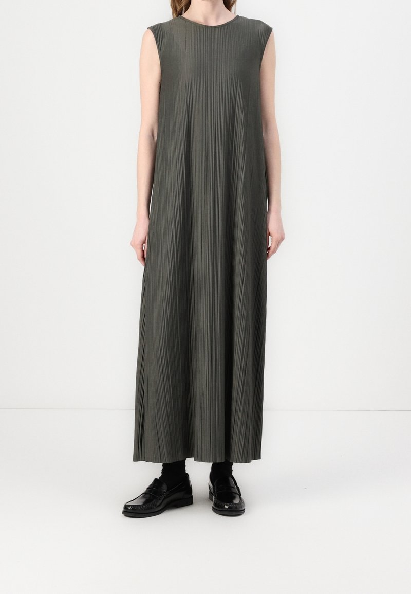 Femme portant une robe maxi plissée sans manches vert olive avec des chaussettes noires et des chaussures habillées noires brillantes, debout sur un fond blanc.