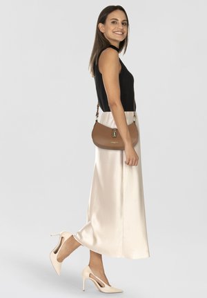 Borsa a tracolla in pelle marrone chiaro con forma curva, hardware dorato e tracolla regolabile. La modella indossa un top nero e una gonna di raso crema.