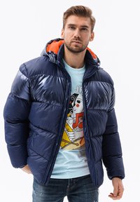 Marineblauwe pufferjack met glanzende textuur, oranje voering in de capuchon, ritssluiting en horizontaal gewatteerd ontwerp, gedragen over een lichtblauw graphic t-shirt.