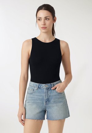 Donna che indossa una canotta nera senza maniche e shorts di denim azzurro chiaro con piccole decorazioni, in piedi contro uno sfondo bianco.