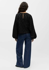 Vrouw met lang krullend haar in een zwarte textuur blouse met lange mouwen en een wijde blauwe jeans, staand met haar gezicht van zich af op een witte achtergrond.
