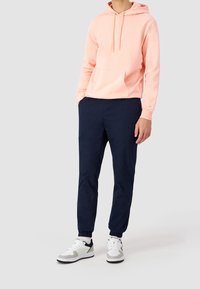 Felpa rosa chiaro con tasca frontale e stringhe, abbinata a pantaloni tapered blu navy e sneakers bianche con dettagli neri.
