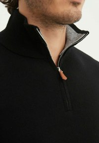 Pull à zip noir avec un col haut, doté d'une doublure intérieure grise et d'une petite garniture en cuir sur la tirette du zip. Texture douce.
