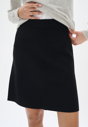 Femme portant une jupe noire au genou et un pull gris clair, ajustant la taille de la jupe avec les deux mains.