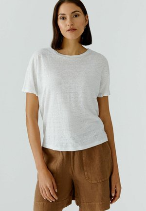 Frau mit schulterlangem dunklem Haar, die ein weißes kurzärmeliges Hemd und braune Shorts trägt, steht vor einem schlichten hellen Hintergrund.