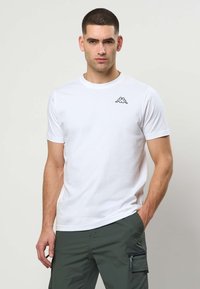 Mand med kort mørkt hår iført en hvid t-shirt med logo og olivengrønne cargobukser, stående med den ene hånd i lommen.