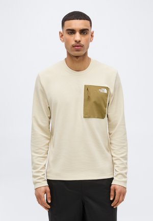 Young man wearing a beige long-sleeve shirt with a green zippered chest pocket and black pants, standing against a plain background.